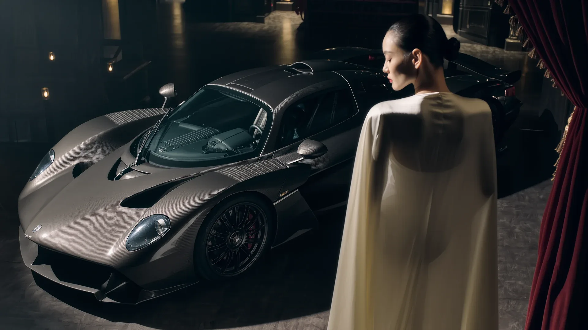 A dark supercar in a theatrical interior with a figure in a long coat beside it