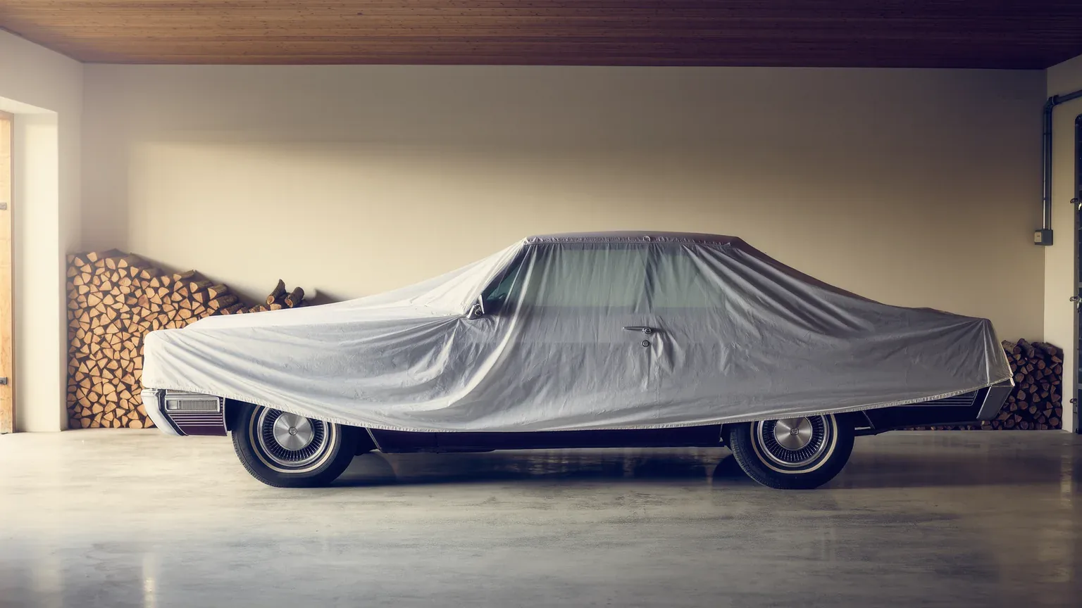 A car under a silver cover in a garage beside a stack of firewood